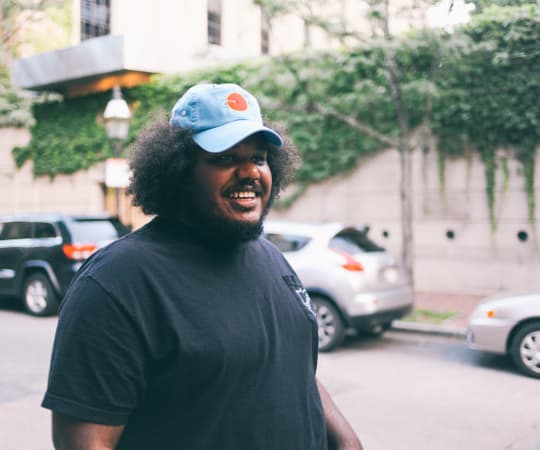 A smiling person with curly hair wearing a black t-shirt and a light blue cap stands on a city street with cars and greenery in the background.