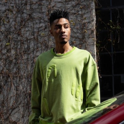 A stylish man wearing a vibrant green sweatshirt with distressed details, standing against a textured wall with vines.