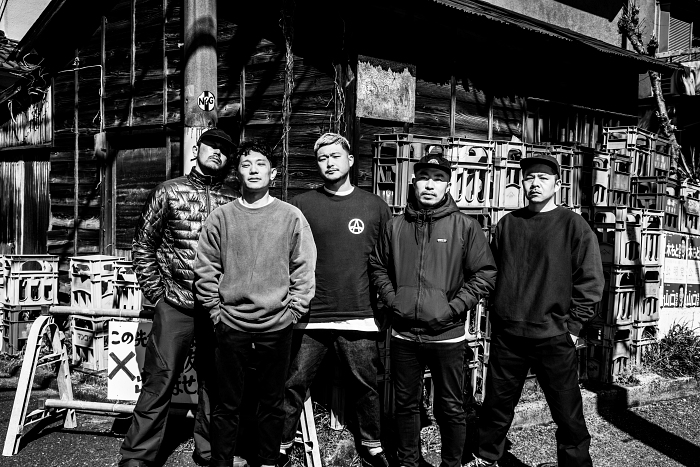 A group of five individuals standing together in front of a rustic wooden building, captured in black and white. They are wearing casual clothing and have varying expressions.