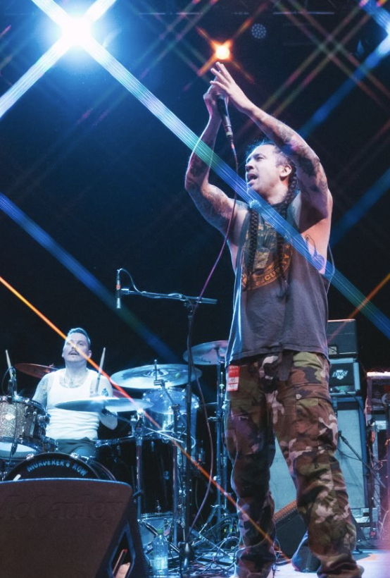 A male vocalist passionately performing on stage with a microphone, while a drummer plays in the background. Colorful stage lights create a vibrant atmosphere.