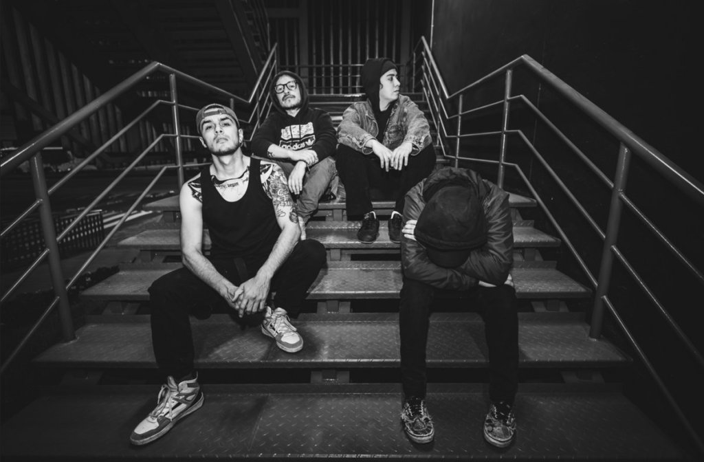 A black and white image of four young men sitting on a staircase. One is wearing a tank top and sneakers, while another is in a hoodie and glasses. Two others are in hooded jackets, with one resting his head on his knees.