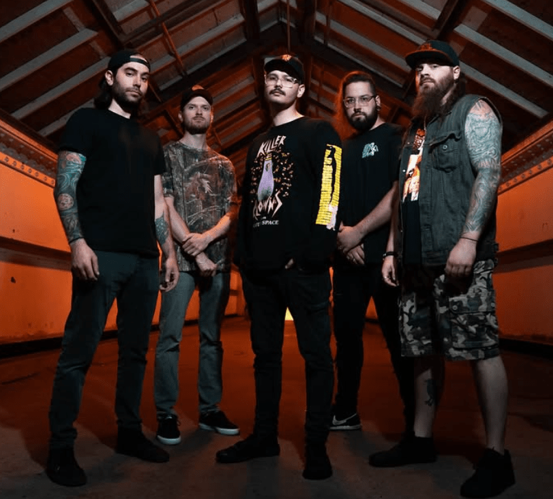 A group of five men standing together in an indoor setting with wooden beams. They are dressed in casual clothing, including graphic t-shirts and jeans, with some sporting tattoos and distinctive facial hair.