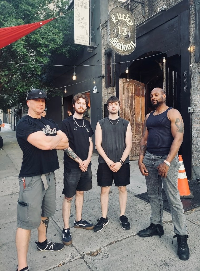Four individuals standing in front of the Lucky 13 Saloon, a bar with a rustic entrance. They are casually dressed, with two men in sleeveless tops and two in t-shirts. The area is outdoors, featuring string lights and a distinct entrance.