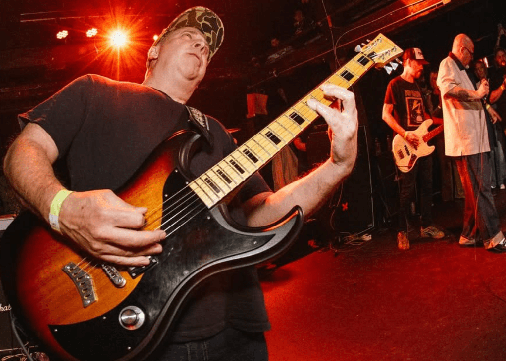 A musician passionately playing an electric guitar on stage with band members in the background.