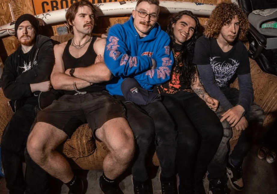 Five young adults sit closely together on a couch, showcasing a casual and relaxed atmosphere. They display a mix of clothing styles, including band t-shirts and casual wear, with some having distinctive hairstyles.
