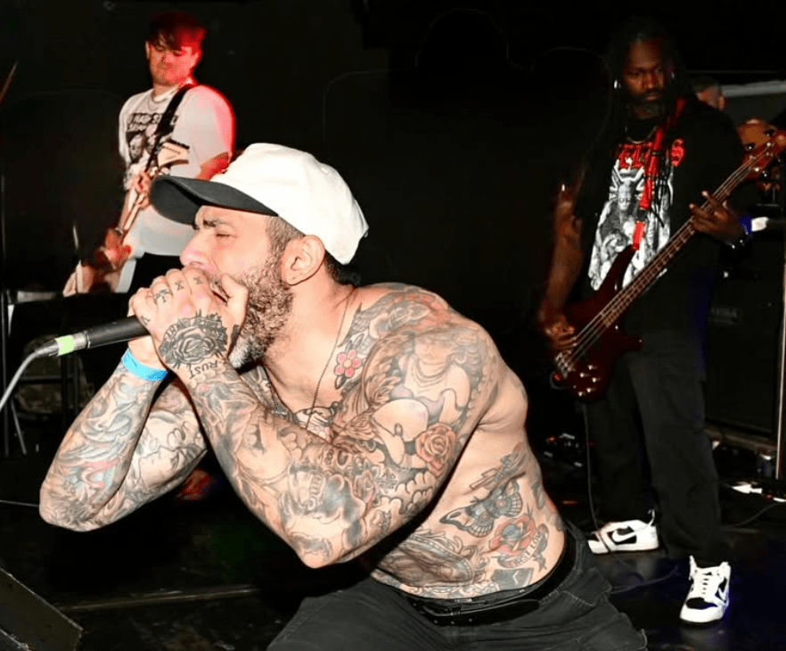 A live music performance featuring a tattooed male vocalist passionately singing into a microphone, with two bandmates in the background playing guitar and bass.