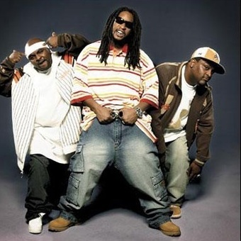 Three men posing together, wearing casual streetwear, with one man in the middle showing confidence. The background is a simple gradient.