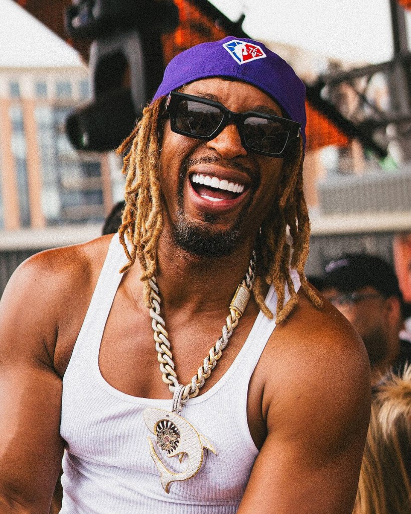 A smiling man with long, curly hair wearing a white tank top, sunglasses, and a purple cap, adorned with a large, decorative necklace.