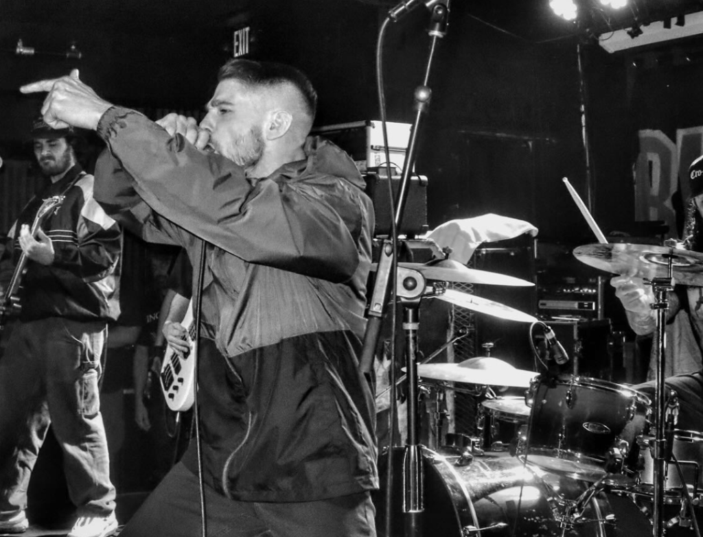 A black and white photo of a live music performance, featuring a male vocalist passionately singing into a microphone while gesturing with his hand. In the background, a guitarist is visible, and a drummer can be seen playing their kit.