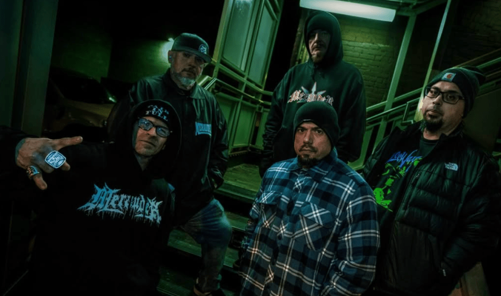 A group of five men posing for a photo in a dimly lit outdoor setting, wearing streetwear and hoodies. The man in the front is making a hand gesture, while the others stand behind him with various expressions.