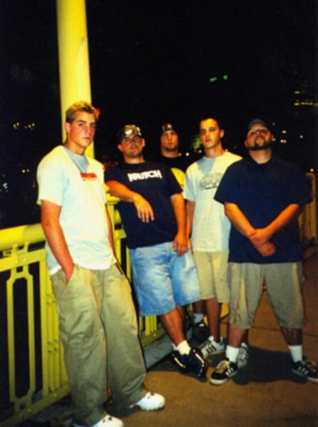 A group of five young men posing together at night on a bridge, wearing casual clothing and standing with relaxed postures.