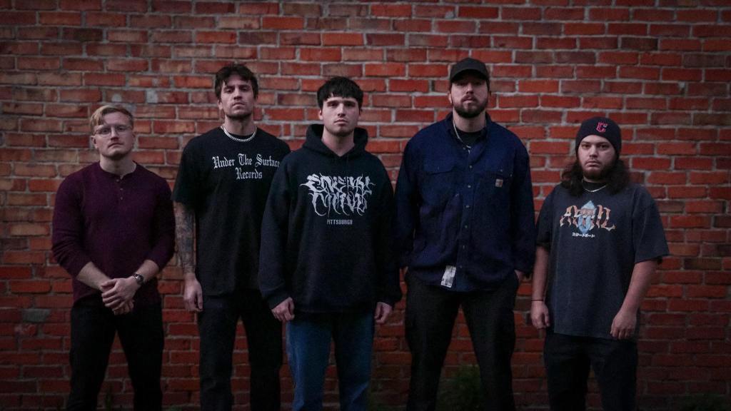 Five men standing in front of a brick wall, wearing casual streetwear, under low light.