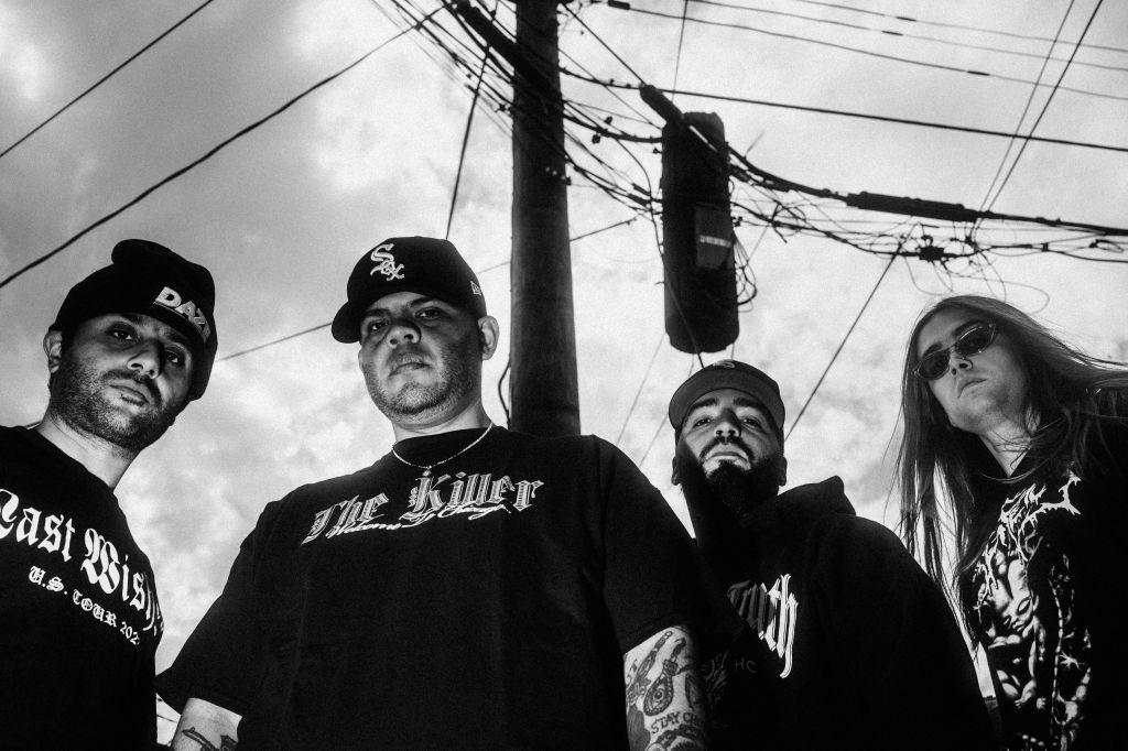 A group of four individuals posing dramatically in front of power lines, wearing black clothing with distinct logos and designs, in a moody black-and-white setting.