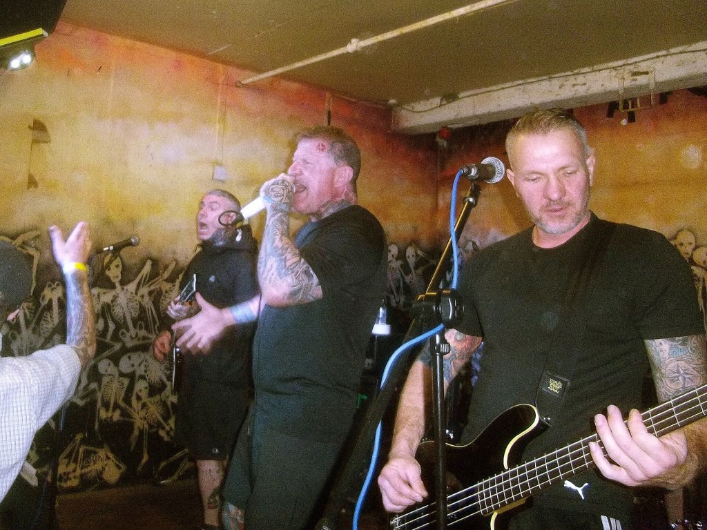 A live music performance featuring three male musicians on stage. One singer is holding a microphone, passionately engaging with the audience, while another musician plays the guitar. The third musician plays bass, appearing focused. The background has a colorful mural with skeletal figures.