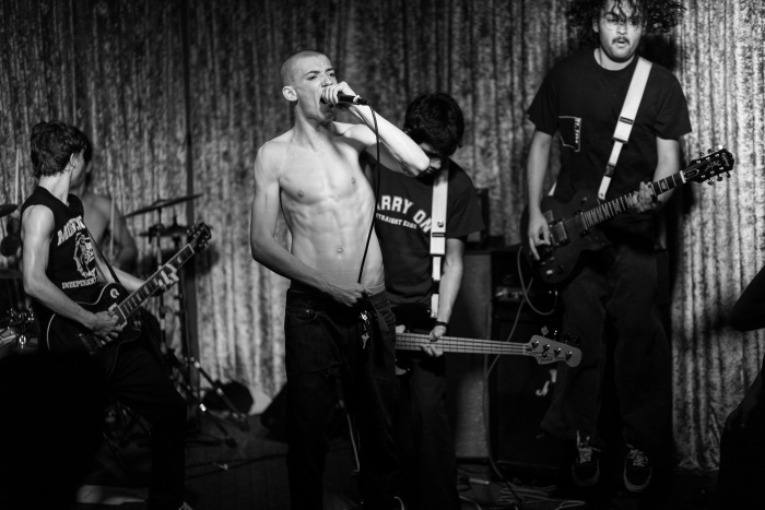 A black and white image of a band performing live on stage. The vocalist, shirtless, passionately sings into a microphone while the other band members play electric guitars and bass. The background features a draped curtain.