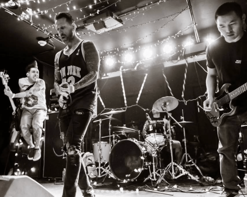 A lively band performing on stage with festive string lights in the background. One musician is jumping enthusiastically while others are focused on their instruments. The scene captures the energy of a live music event.