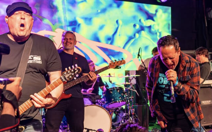 A band performing live on stage with vibrant colorful visuals in the background, featuring a guitarist passionately playing and a vocalist energetically singing into a microphone.