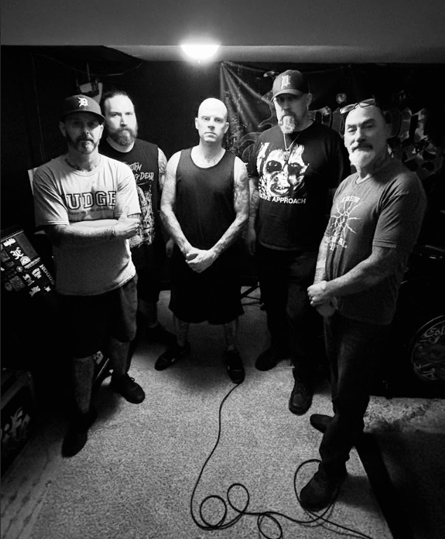 Five band members posing together in a music rehearsal space, wearing concert T-shirts and displaying tattoos. The scene is in black and white with low lighting.