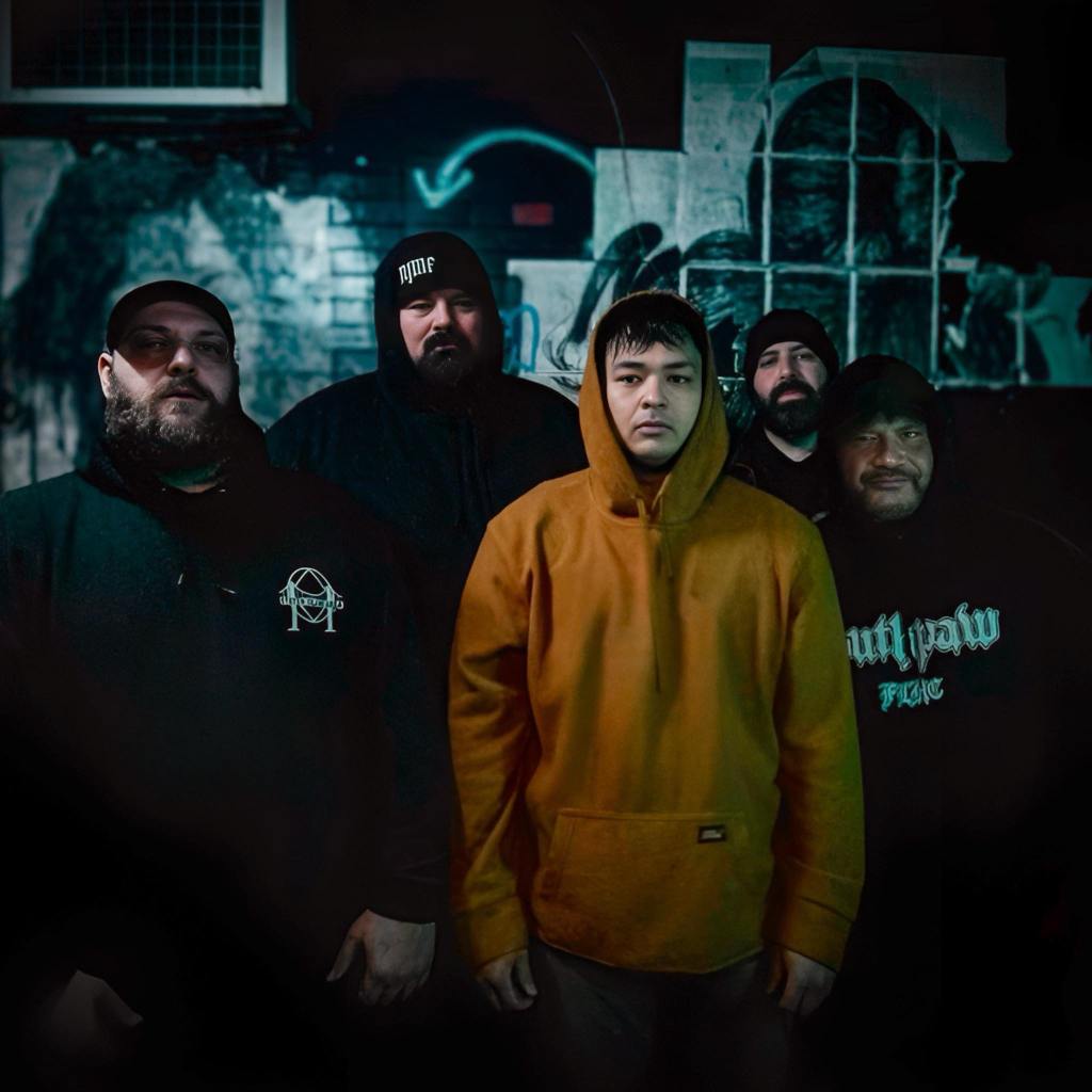 A group of five men standing together in front of a graffiti-covered wall, dressed in dark hoodies and beanies, with one wearing a bright yellow hoodie.