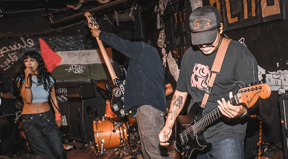 A live band performance featuring a female vocalist and two male musicians, one playing an electric guitar and the other playing bass. The stage has a vibrant atmosphere with graffiti and a Palestinian flag in the background.