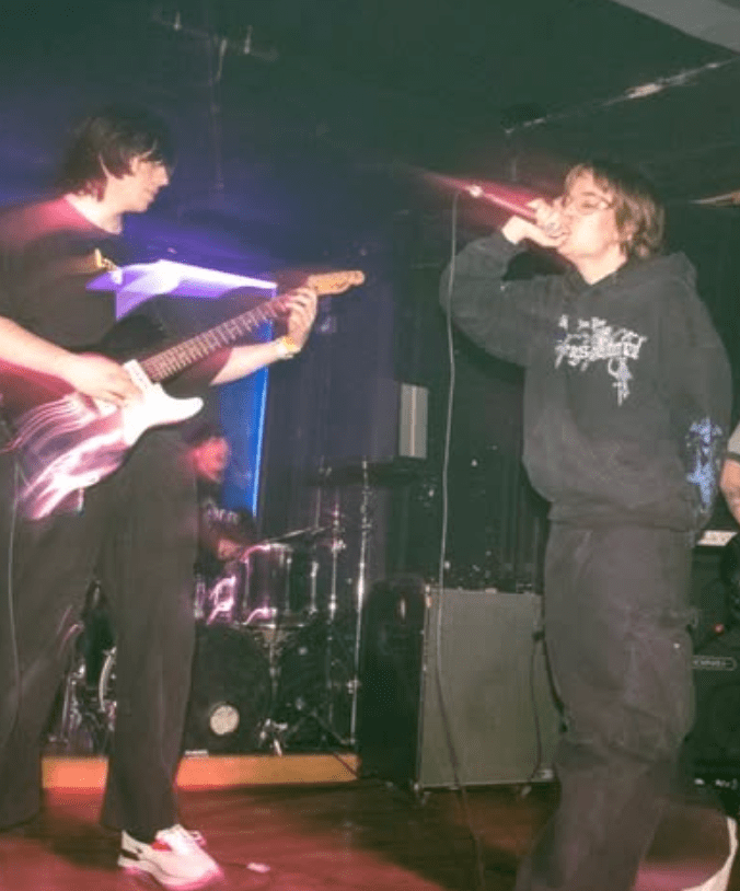 Two musicians performing on stage, one playing an electric guitar and the other singing into a microphone. The setting is dimly lit with colorful stage lighting.