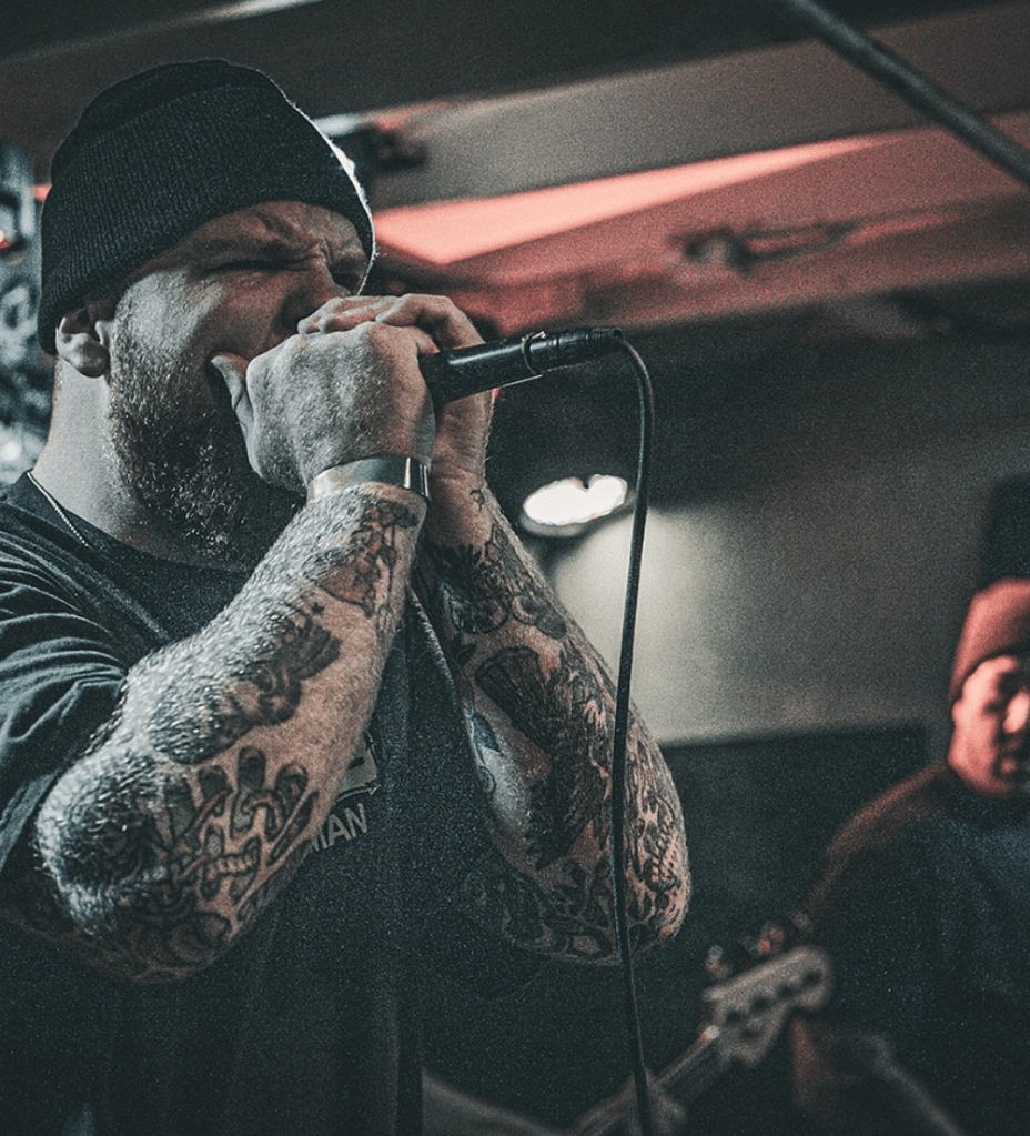 A tattooed vocalist passionately singing into a microphone on stage, wearing a black beanie and a dark shirt, with a bassist partially visible in the background.
