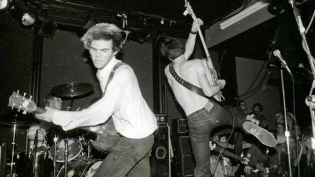Black and white image of a rock band performing live on stage, with two guitarists prominently featured in dynamic poses.