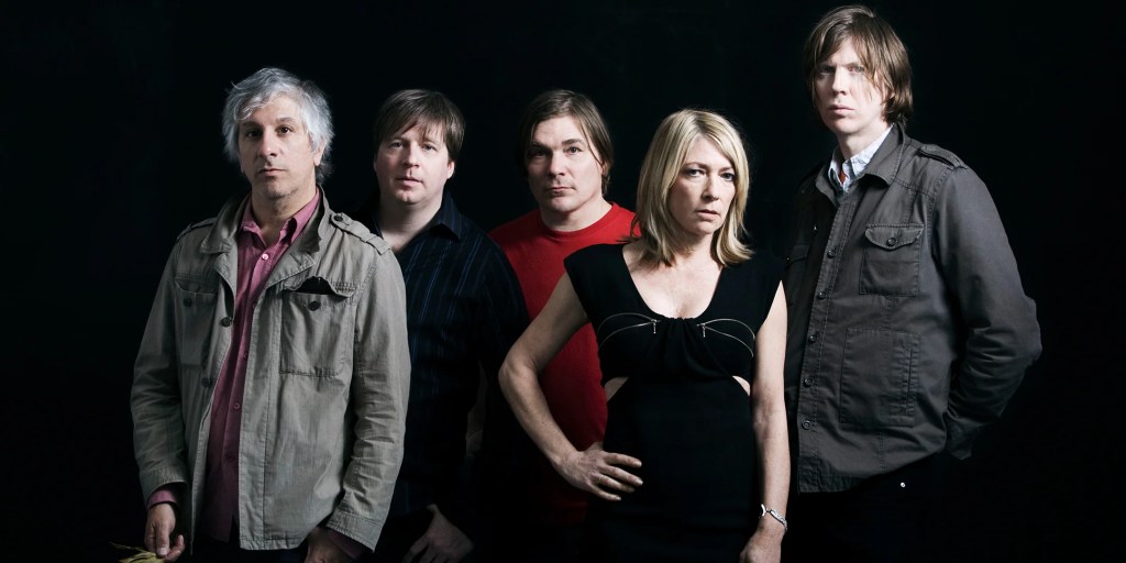 Five musicians posed against a dark background, wearing casual clothing and displaying a serious demeanor.