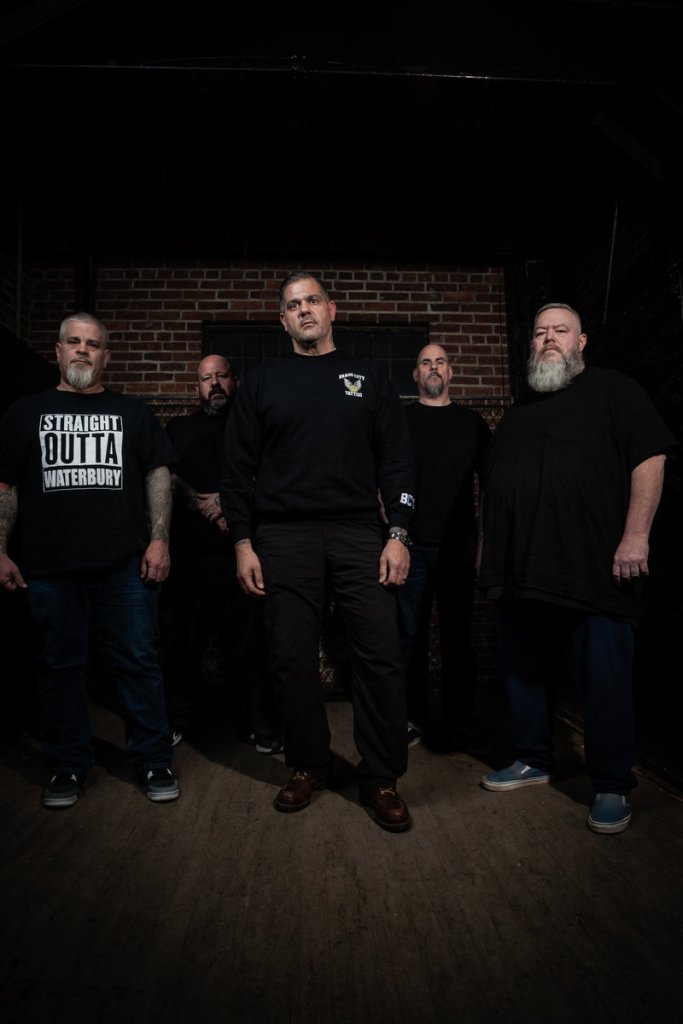 Five men standing together in a dimly lit room with a brick wall in the background, showcasing a mix of casual and streetwear styles.