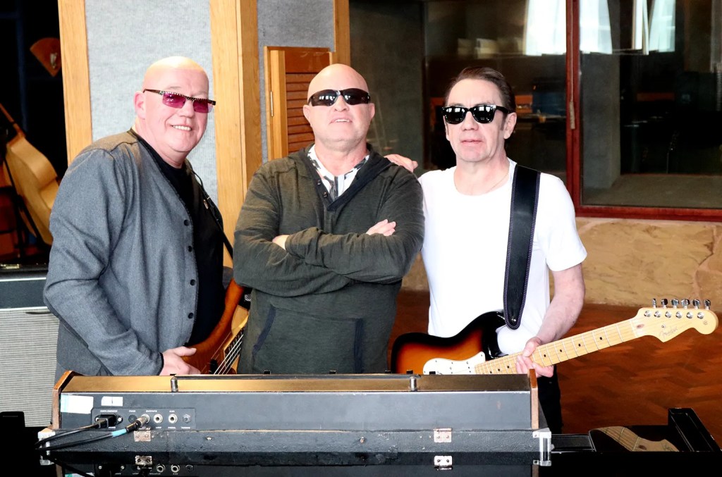 Three musicians posing with instruments in a recording studio, wearing sunglasses, with a keyboard in the foreground.