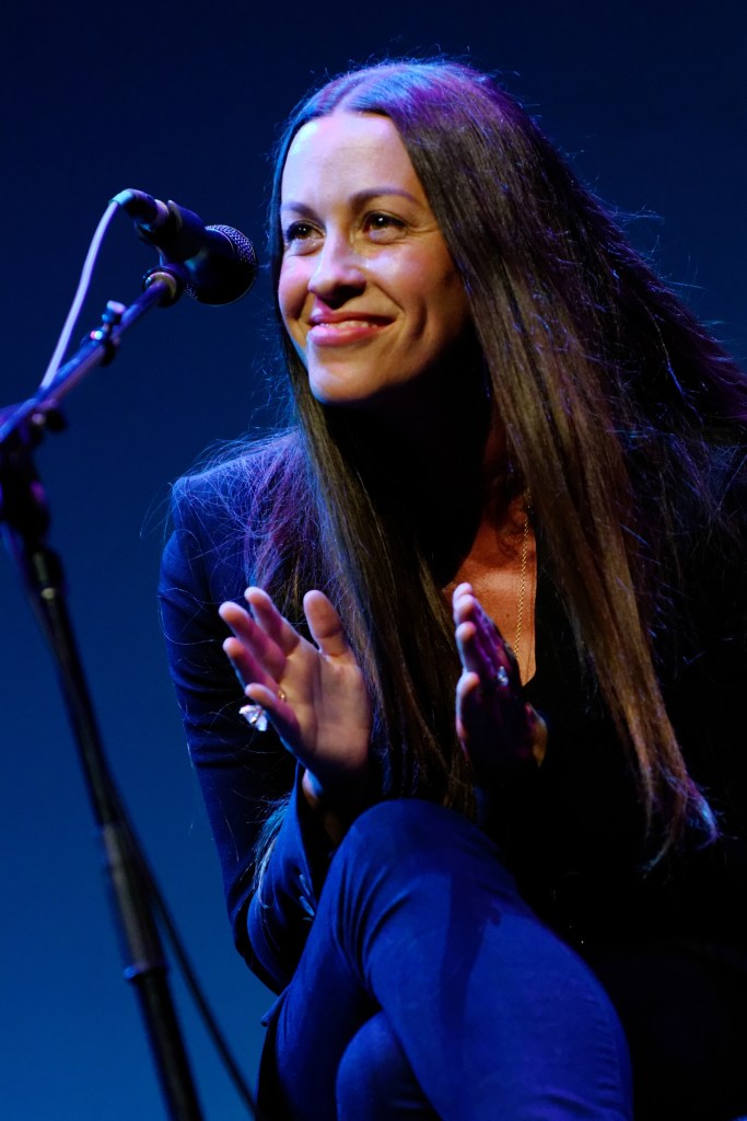 A woman with long, dark hair smiles while sitting on a stool beside a microphone, clapping her hands in a performance setting.