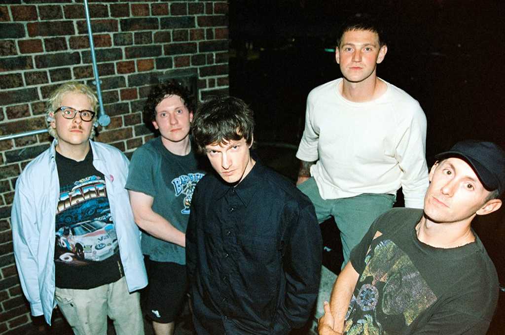 A group of five young men standing against a brick wall, with a mix of casual clothing styles. The lighting is dim, adding a moody atmosphere to the scene.