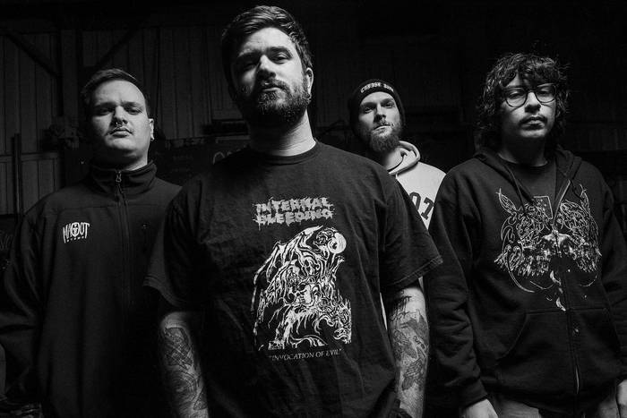 A black and white photo of four men standing together, dressed in band T-shirts and hoodies, with serious expressions. The background appears dimly lit, emphasizing their presence.