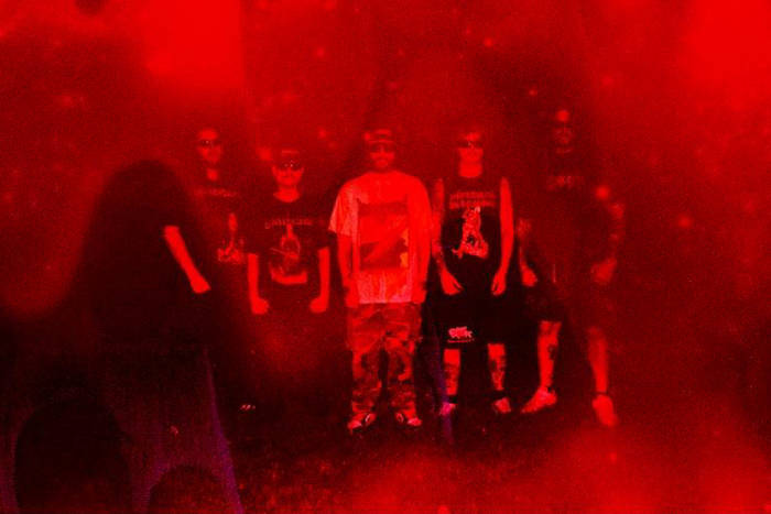A group of six individuals dressed in dark clothing, some wearing sunglasses, standing together against a vibrant red background.