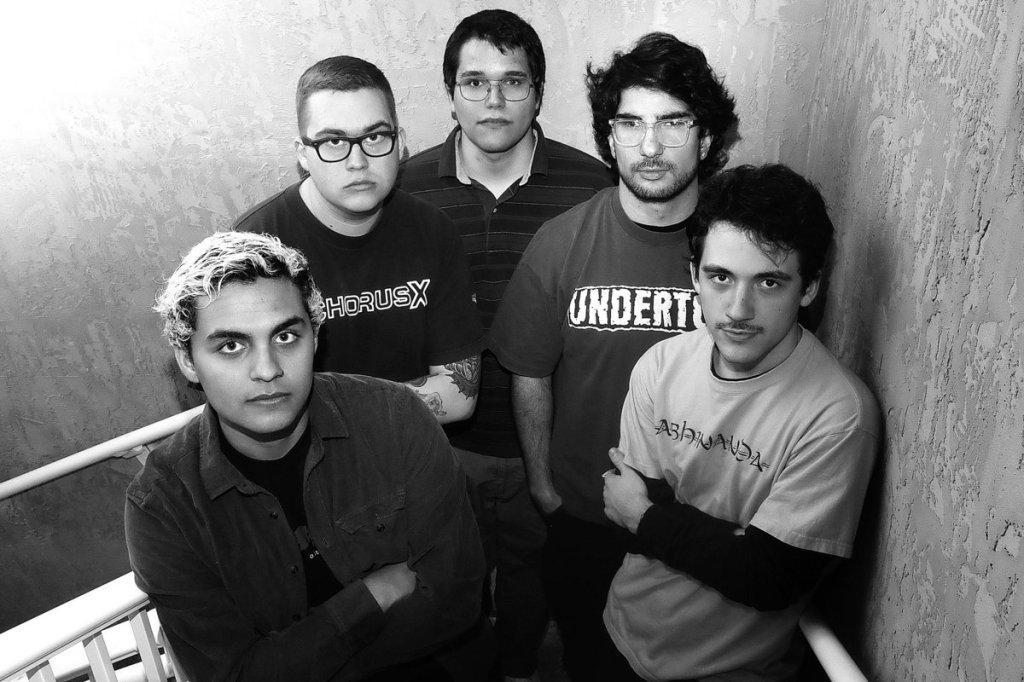 A black and white image of five individuals standing on a staircase, looking directly at the camera. They are dressed in casual clothing and display a variety of hairstyles.