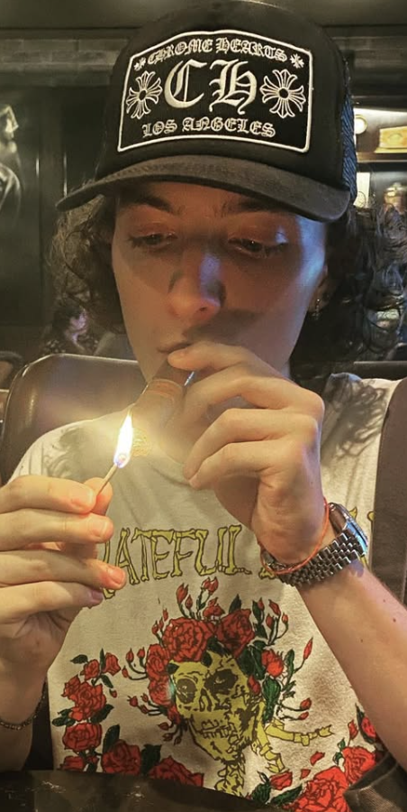 A person with curly hair wearing a black cap is lighting a small object with a lighter while sitting at a table. They are dressed in a white t-shirt featuring a colorful design with roses.