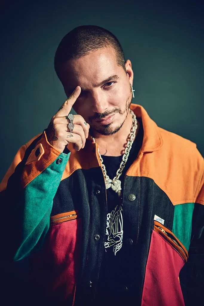 A close-up portrait of a man with a short haircut, wearing a colorful jacket with orange, green, and black sections, and silver jewelry. He is posing with one hand raised to his forehead.