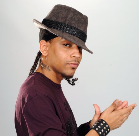 A person wearing a dark fedora hat, with braided hair and a wristband, clapping hands while looking at the camera.