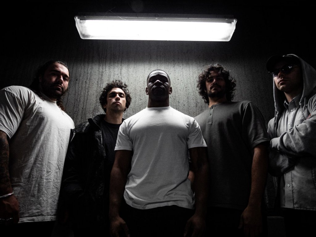 A group of five men standing in a dimly lit setting, illuminated by an overhead light. They display a serious demeanor, dressed in casual clothing.