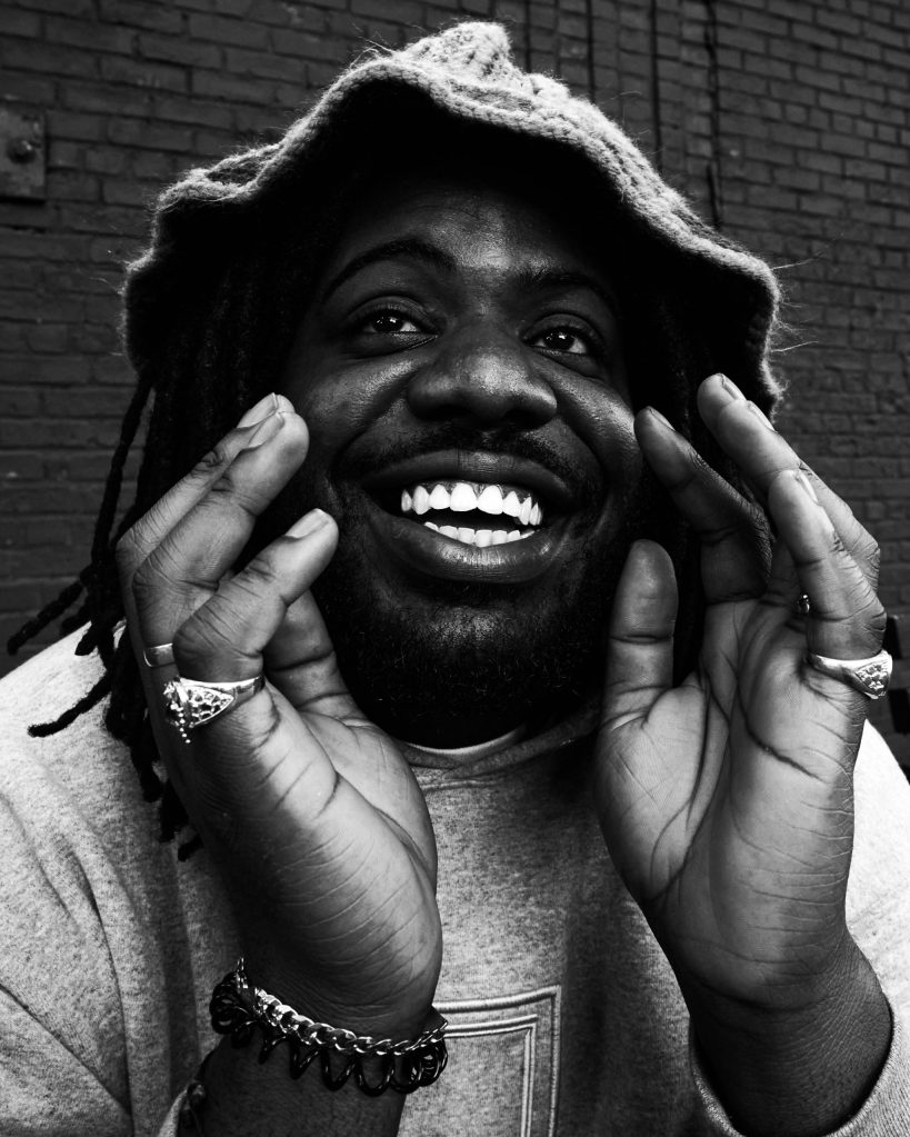 A close-up portrait of a smiling person with curly hair wearing a hoodie, gesturing with hands near their face in an expressive manner. The image is in black and white.