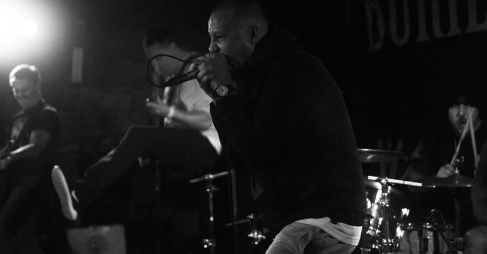 A live music performance featuring a vocalist passionately singing into a microphone, while a guitarist and drummer play in the background. The image is in black and white and captures the energetic atmosphere of the concert.