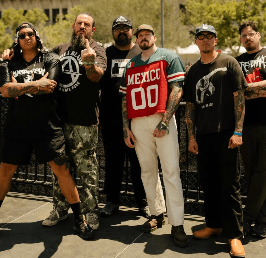 A group of seven tattooed individuals posing together outdoors, wearing casual and band-themed clothing, with some showing tattoos and making various expressions.