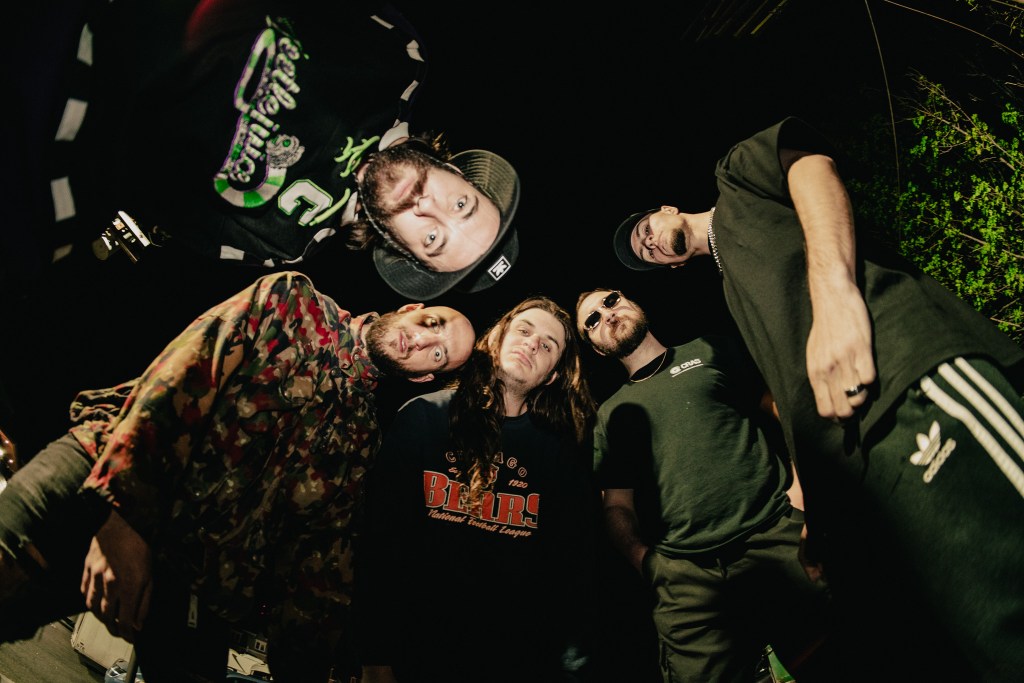A group of five individuals posing together in a low angle shot, with various styles of clothing and accessories, under dim lighting.