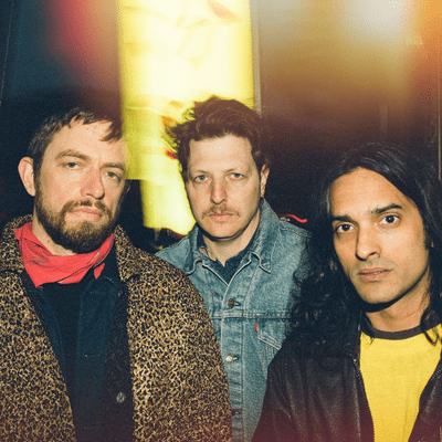 Three men posing for a photo, with one wearing a leopard print coat and red scarf, another in a denim jacket, and the third in a yellow shirt, set against a colorful background.