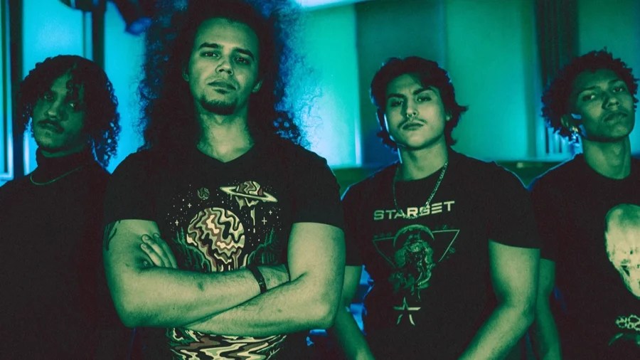 A group of four young men standing together in a dimly lit room with a blue-green hue, dressed in casual attire featuring graphic t-shirts. They strike confident poses, with one individual in the foreground crossing his arms.