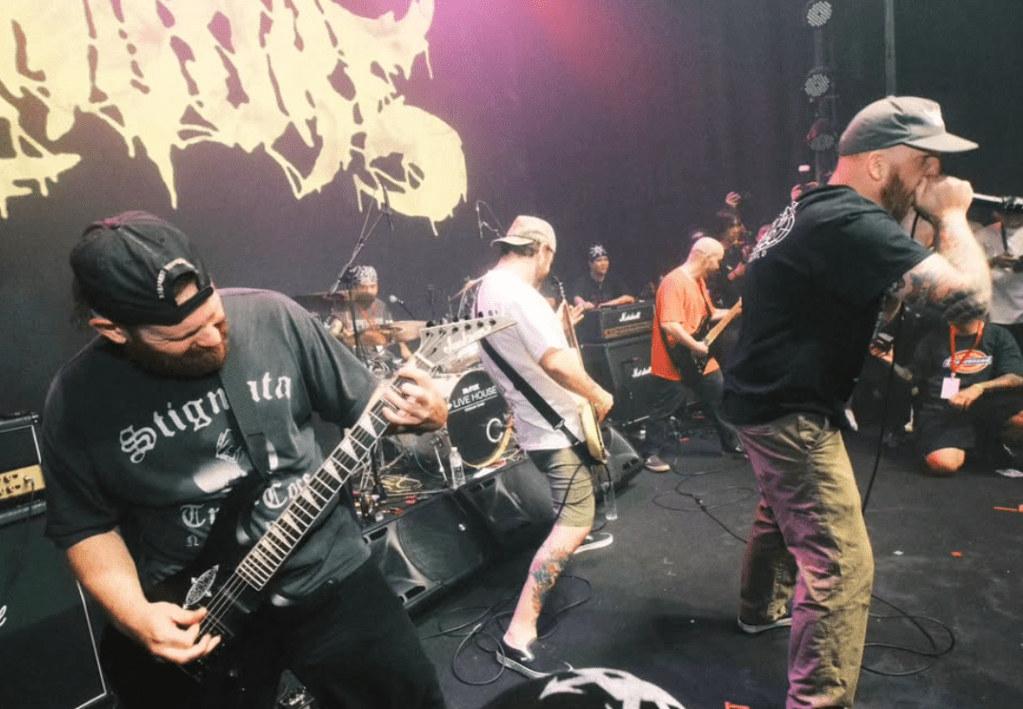 A live music performance featuring multiple musicians on stage, with a focus on a guitarist playing an electric guitar and a vocalist singing passionately. The background includes amplifiers and a backdrop with stylized text.
