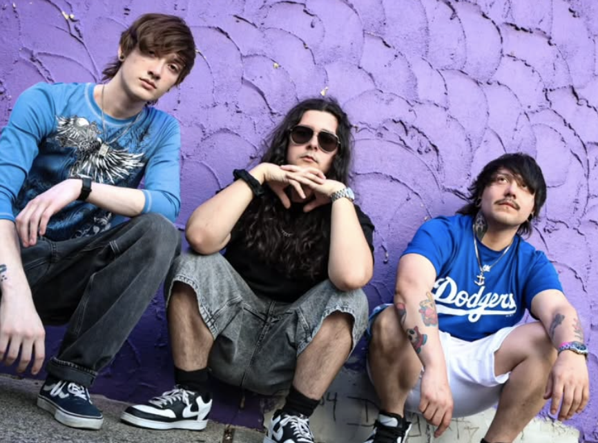 Three young men sitting in front of a purple textured wall, dressed in casual streetwear, with tattoos and different hairstyles.
