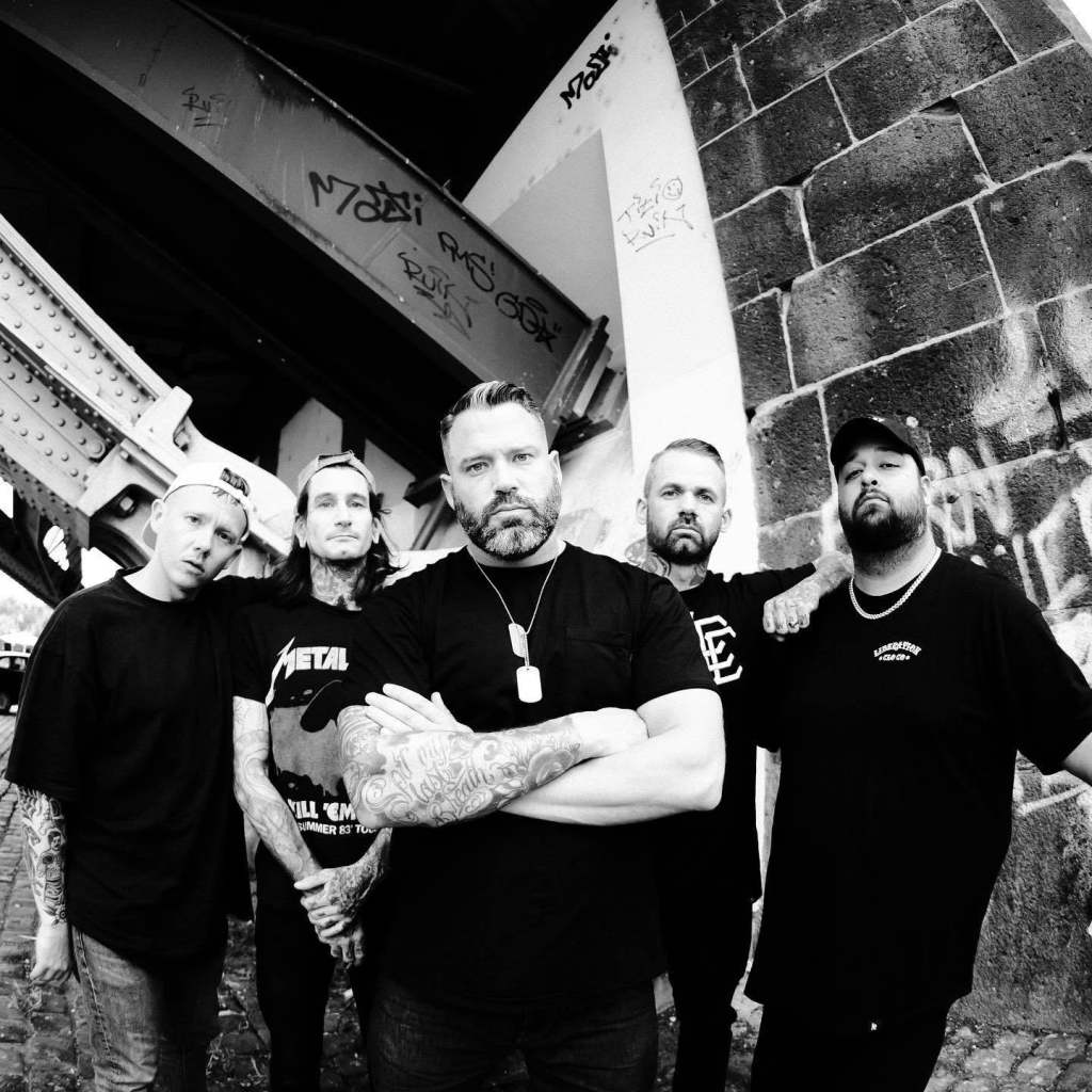 A group of six men posing together under an urban bridge, with graffiti visible in the background, dressed in casual black clothing and showcasing various tattoos.