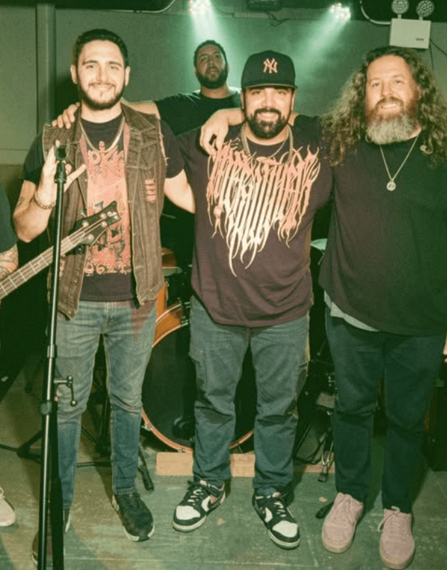 A group of four musicians standing together on stage, smiling and posing for a photo. They are wearing various band T-shirts and casual clothing, with instruments visible in the background, including a guitar and drums.