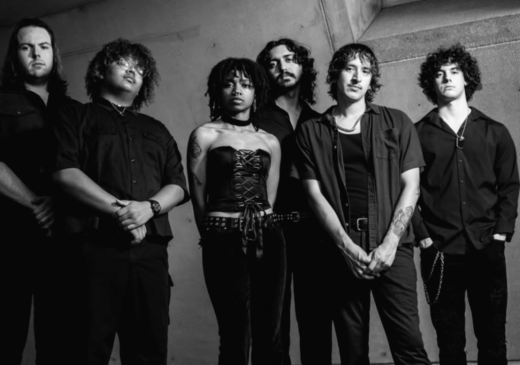 A black and white image of a six-member band standing together, dressed in dark clothing, with various hairstyles and accessories, posed in front of a concrete backdrop.